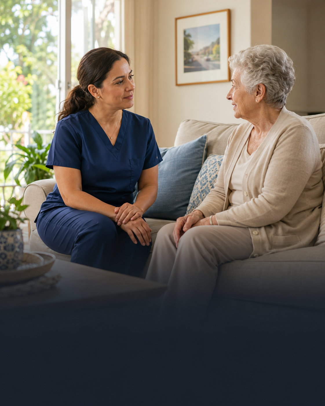 A home health nurse in navy scrubs sitting on a sofa with an older woman, talking with her.