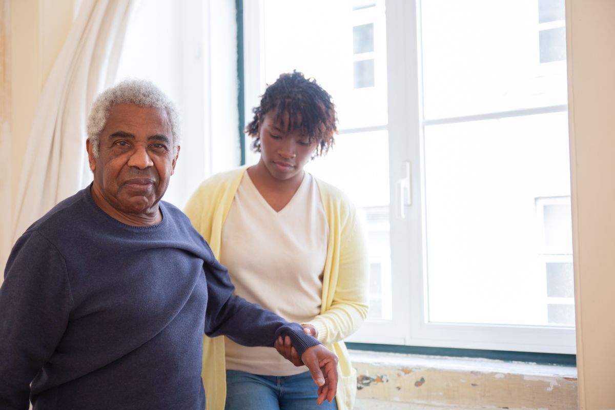 A care professional steadying an older man by the arm near a bright window