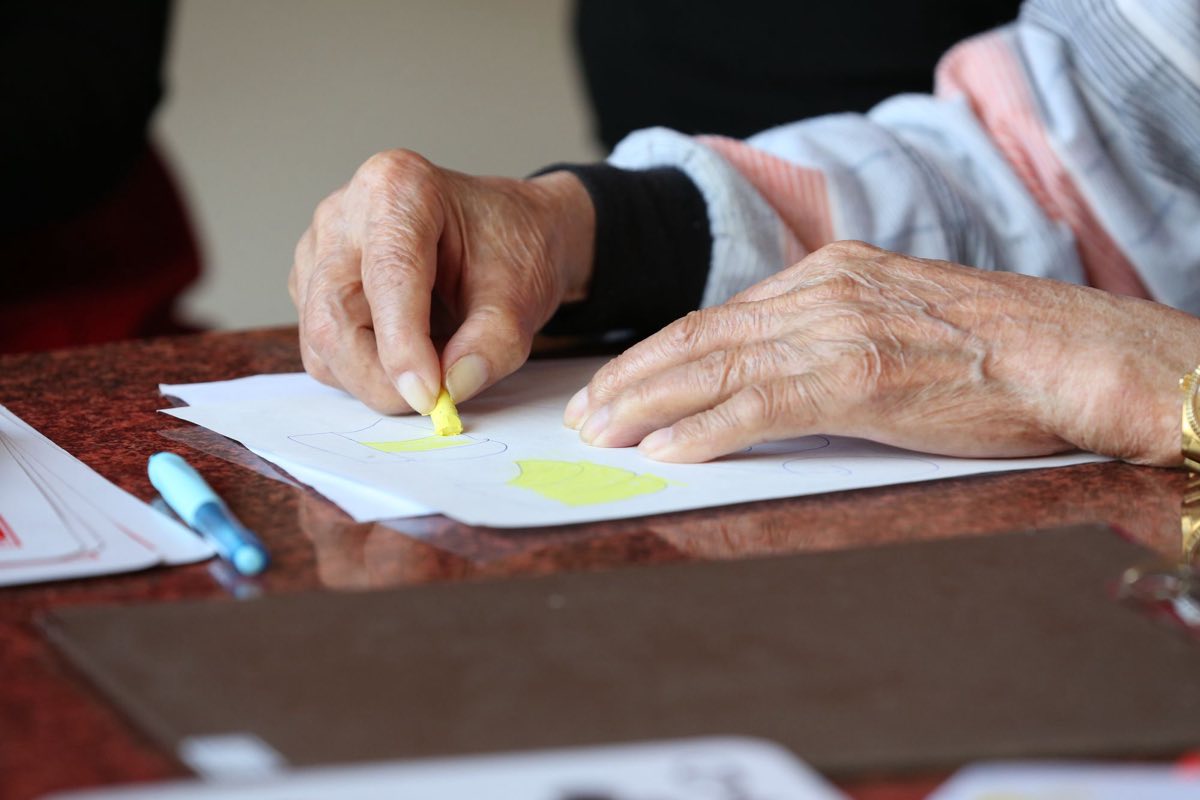 An older adult’s hands using a coloured pencil to complete a fine-motor worksheet at a table
