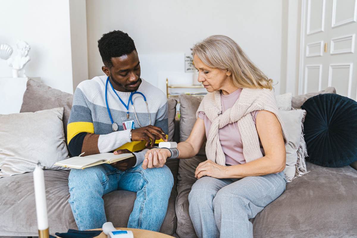 A nurse with a stethoscope and notes taking an older woman’s blood pressure in her living room